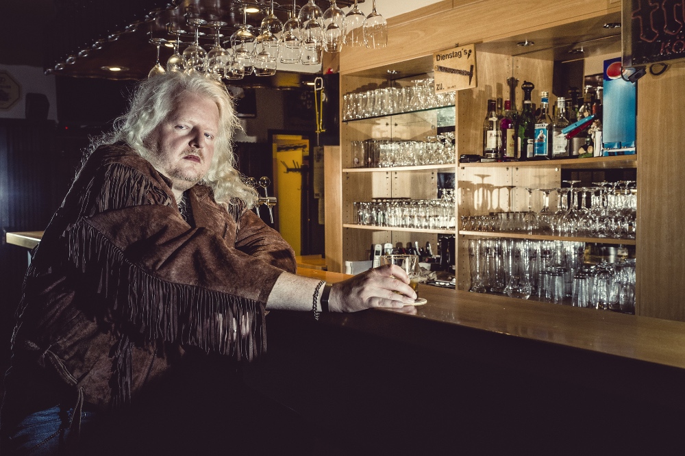 Ein blonder Rocker in Lederjacke sitzt in einer Kneipe an der Theke.