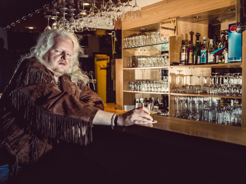 Ein blonder Rocker in Lederjacke sitzt in einer Kneipe an der Theke.