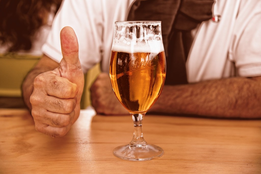 Ein Mann sitzt vor einem Glas Bier und zeigt den Daumen nach oben.