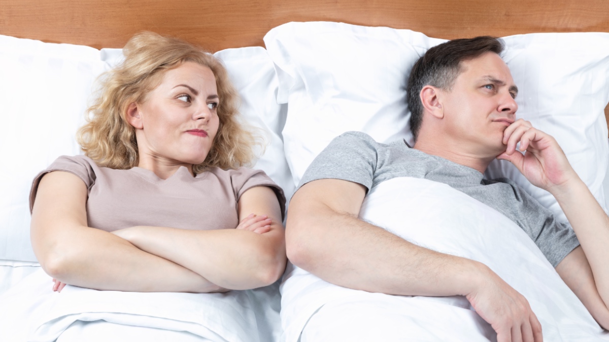 Zwei im Bett, eine Frau blickt misstrauisch auf einen Mann, der sich in die andere Richtung gedreht hat.