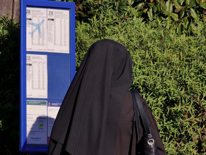 Eine Nonne steht an einer Bushaltestelle vor einem Fahrplan.