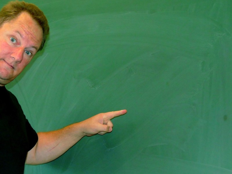 Ein Lehrer zeigt im Klassenzimmer mit dem Finger auf eine leere Tafel.